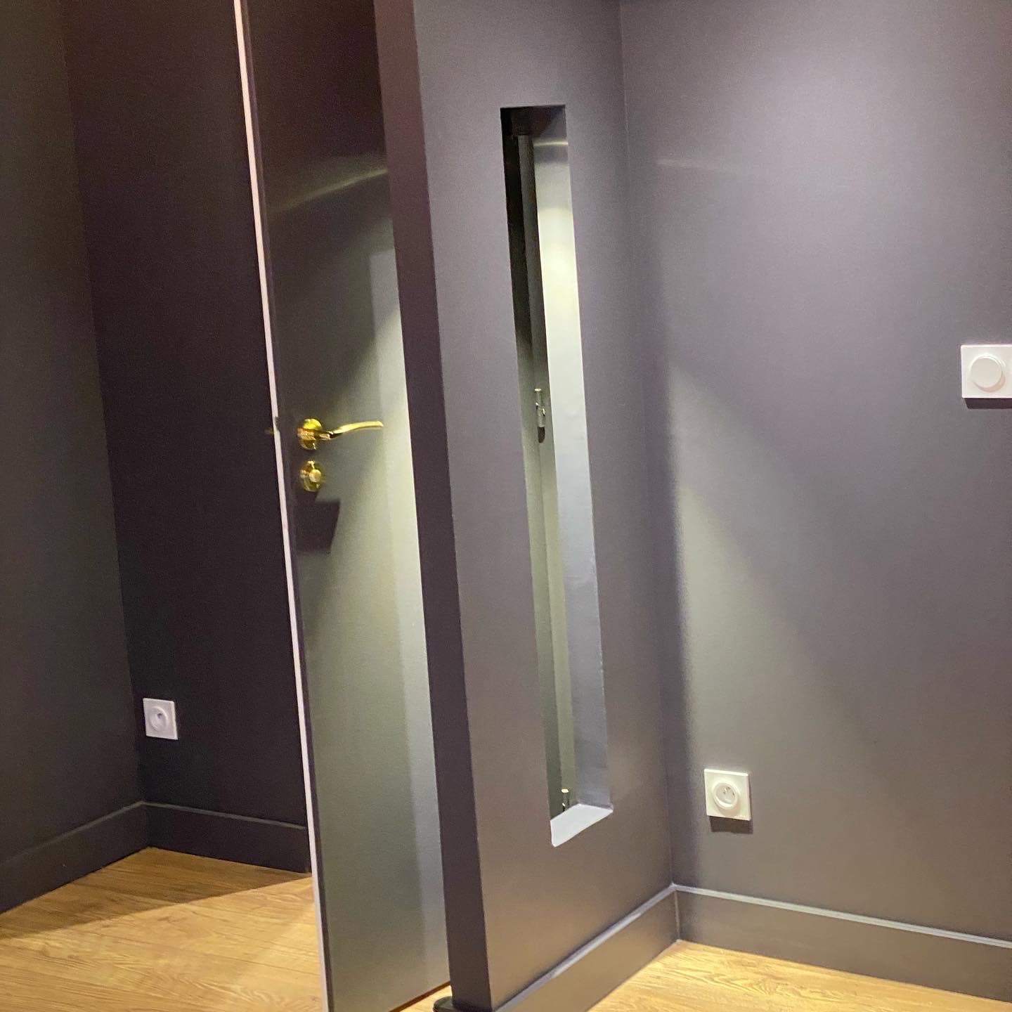 Cloison peinte anthracite percée d’une ouverture verticale étroite, porte assortie à poignée dorée, studio rénové à Lyon