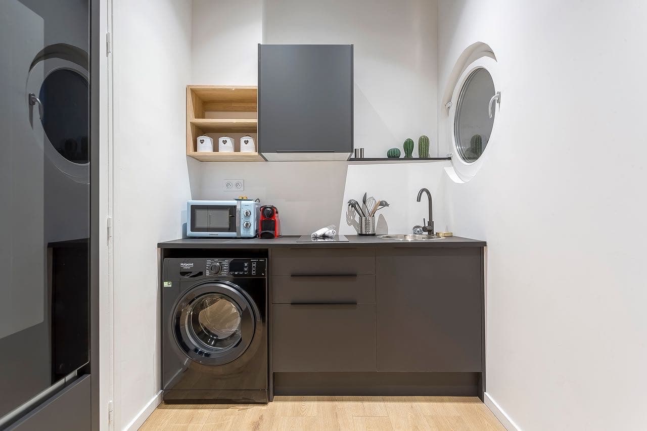 Kitchenette gris anthracite avec lave-linge sous plan, étagère en bois clair et fenêtre ronde à cadre blanc, studio à Bourgoin-Jallieu