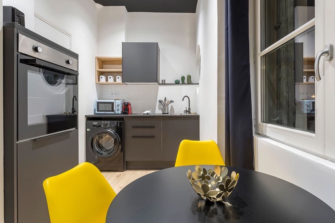 Kitchenette grise avec four en colonne et lave-linge intégré, table ronde noire et chaises jaunes, plafond noir, Bourgoin-Jallieu