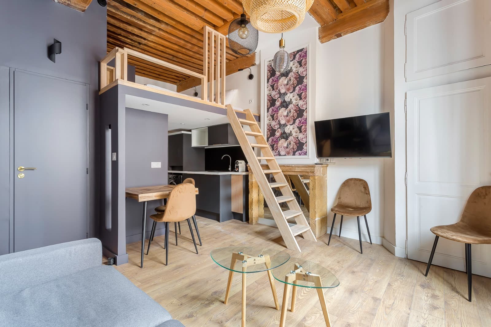 Studio meublé à Lyon : mezzanine à garde-corps bois au-dessus d’une cuisine anthracite, solives apparentes et papier peint floral